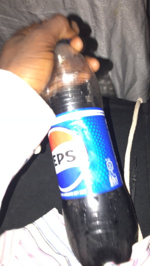 Dark-skinned hand holds Pepsi bottle, white shirt sleeve, gray bed.