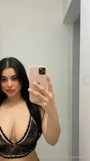 Woman with long black hair in black lace bra taking mirror selfie indoors.