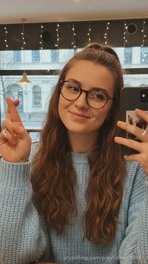 Woman with long brown hair and glasses wearing a light blue sweater, taking a mirror selfie indoors.