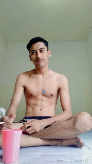 Shirtless man with defined abs sitting cross-legged on floor, wearing dark shorts, holding phone.