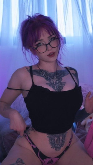 Woman with purple hair and tattoos wears black top and pink underwear, sitting indoors.