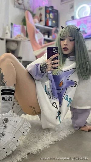 Woman with green hair and tattoos in oversized hoodie, taking mirror selfie in room with pink decor.
