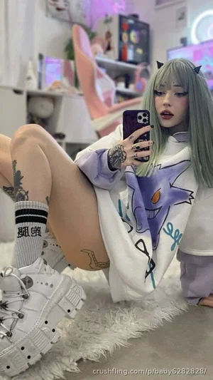 Woman with green wig and tattoos wears oversized hoodie and white boots, taking mirror selfie in
