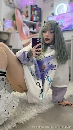 Woman with green hair and tattoos in oversized hoodie, taking mirror selfie in bedroom.