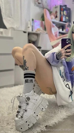 Person with green hair and tattoos in a white hoodie and socks, sitting on a fluffy rug in a