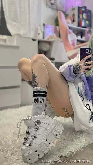 Person with green hair and tattoos wearing socks and white boots, sitting on a fluffy rug in a