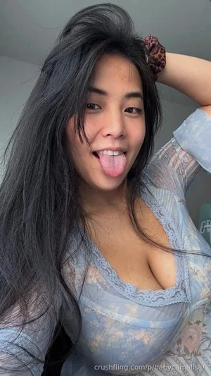 Woman with long dark hair in a sheer blue lace top, posing indoors with tongue out.