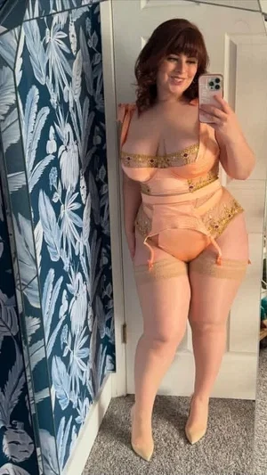 Woman in peach lingerie with gold trim takes mirror selfie in bedroom.