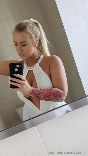 Blonde woman in white halter top with floral tattoo on arm, taking mirror selfie in bathroom.