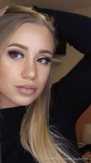 Blonde woman in black top, posing indoors with hand in hair.