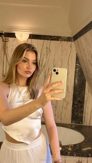 Woman with blonde hair in white crop top and skirt taking mirror selfie in marble bathroom.