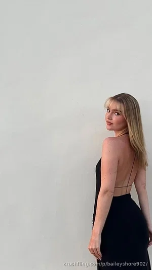 Woman with blonde hair in black backless dress, standing against white wall.