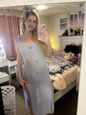 Woman in light blue dress with butterfly details takes mirror selfie in bedroom.