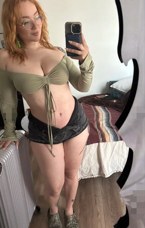 Redhead woman in green top and shorts taking mirror selfie in bedroom.