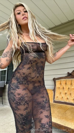 Woman with long blonde hair in black lace bodysuit, standing outdoors near a house.