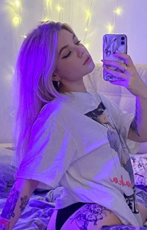Blonde woman in t-shirt taking selfie on bed with purple lights.