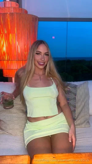 Blonde woman in lime green crop top and mini skirt, sitting on outdoor sofa with drink, evening sky