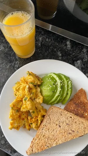 Plate with scrambled eggs, avocado slices, toast, and a glass of orange juice on a dark countertop.
