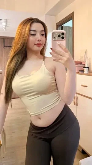 Woman with long hair in beige crop top and black leggings taking mirror selfie in bedroom.