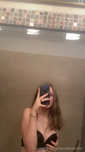 Woman in black bra taking mirror selfie in tiled bathroom.