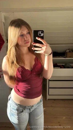 Woman with blonde hair in red lace top and jeans, taking mirror selfie in bedroom.
