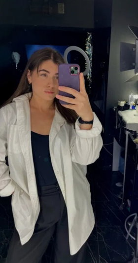 Woman with long brown hair in white shirt taking mirror selfie indoors.