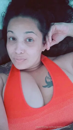 Woman with dark curly hair in orange top, lying down, chest tattoo visible.