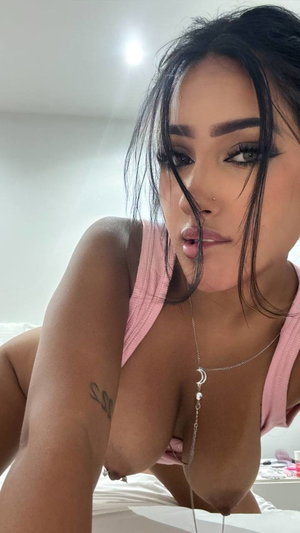 Woman with dark hair in pink top posing on bed, wearing necklace, tattoo on arm.