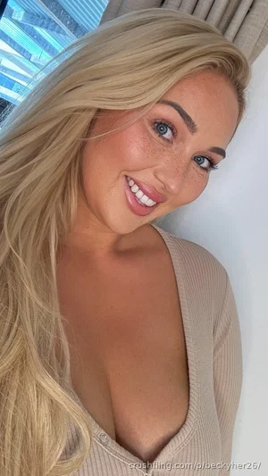 Blonde woman in a beige ribbed top, smiling, indoors near a window.