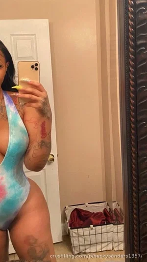 Woman in tie-dye one-piece swimsuit with tattoos, taking mirror selfie in bedroom.