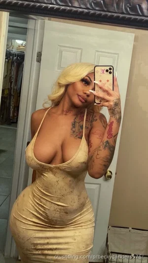 Woman with blonde hair and tattoos wears a tight beige dress, taking a mirror selfie in a bedroom.