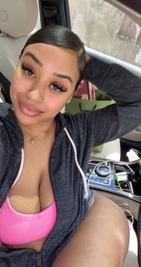 Woman in pink top and gray hoodie sitting in car, smiling at camera.