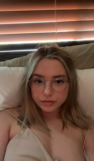 Woman with blonde hair and glasses lying in bed, wearing a light-colored top.