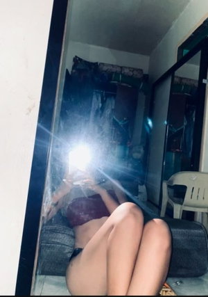 Woman in maroon top and dark pants lying on couch, taking mirror selfie in dim room.