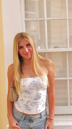 Blonde woman in floral top and jeans standing by a window.