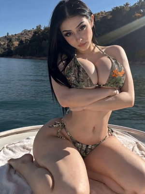 Woman in camouflage bikini posing on boat by water, arms crossed.