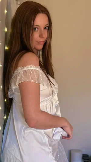 Woman with long brown hair in white lace-trimmed nightgown, standing indoors near string lights.