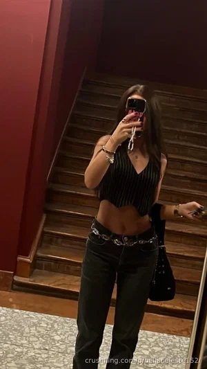 Woman in black striped crop top and jeans, holding phone, standing by wooden stairs indoors.