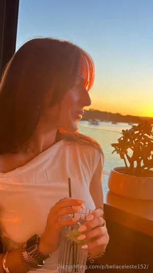 Woman with long dark hair in white off-shoulder top holding drink by sunset waterfront.