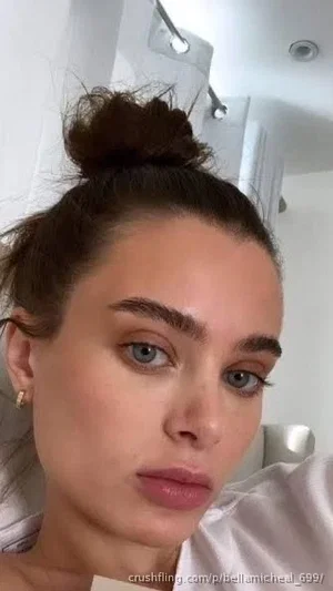 Woman with brown hair in bun, wearing white top, indoors, close-up selfie.