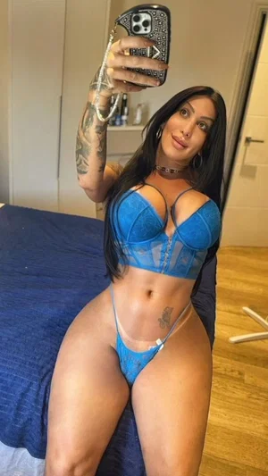 Woman in blue lace lingerie poses on bed, tattoos visible, holding phone for mirror selfie.