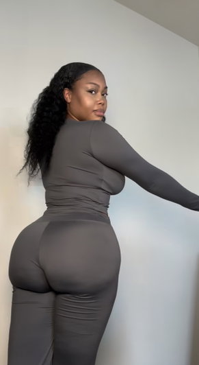 Curvy woman in tight grey outfit posing indoors from behind.