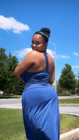 Woman in blue dress posing outdoors on a sunny day, back to camera, smiling over shoulder.