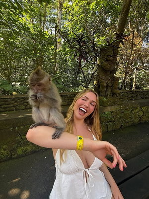 Woman in white dress with monkey on shoulder, smiling in lush green outdoor setting.