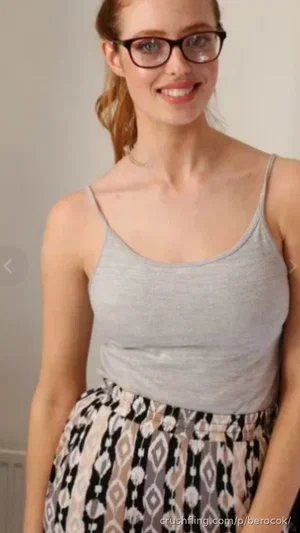 Woman with glasses in gray tank top and patterned skirt, smiling indoors.
