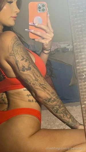 Woman in orange lingerie with tattoos, taking mirror selfie in bedroom.
