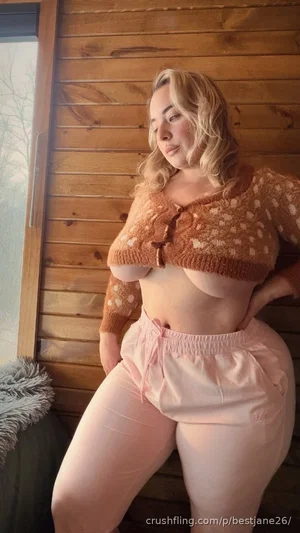 Woman with blonde hair in a brown knitted crop top and pink pants, posing indoors against wood