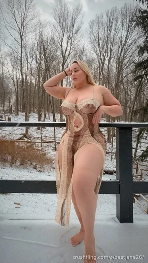 Woman in beige lace corset standing barefoot on snowy deck, surrounded by leafless trees.