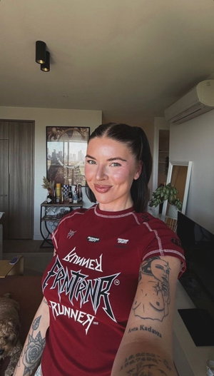 Woman with tattoos in red graphic tee, smiling in indoor living room setting.