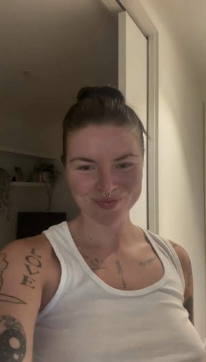 Woman with tattoos in white tank top, indoor setting, smiling at camera.